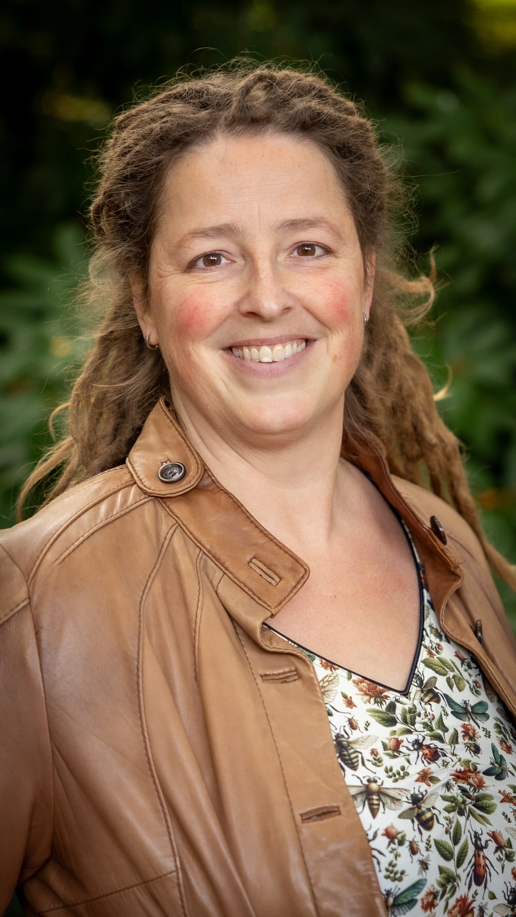 Inge Vervoorn lacht in de camera, in een leren jas voor een groene struik