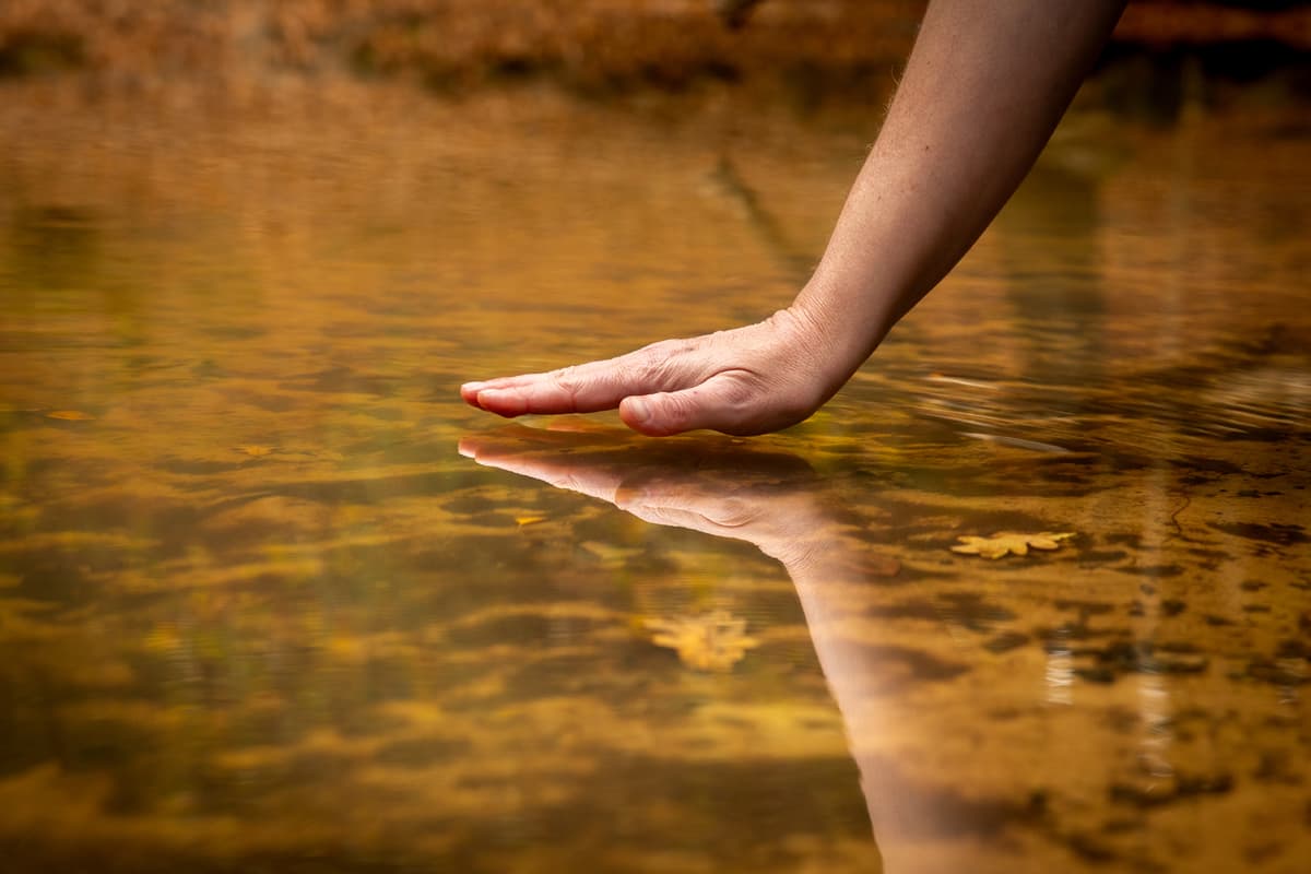 Een hand raakt het stille, goudkleurige water en zijn eigen spiegelbeeld