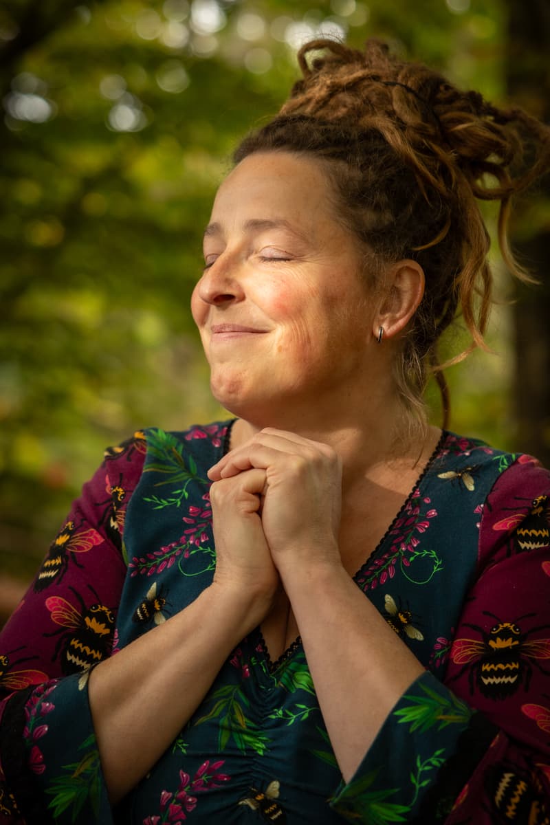 Inge staat met gesloten ogen en gevouwen handen te glimlachen in het bos